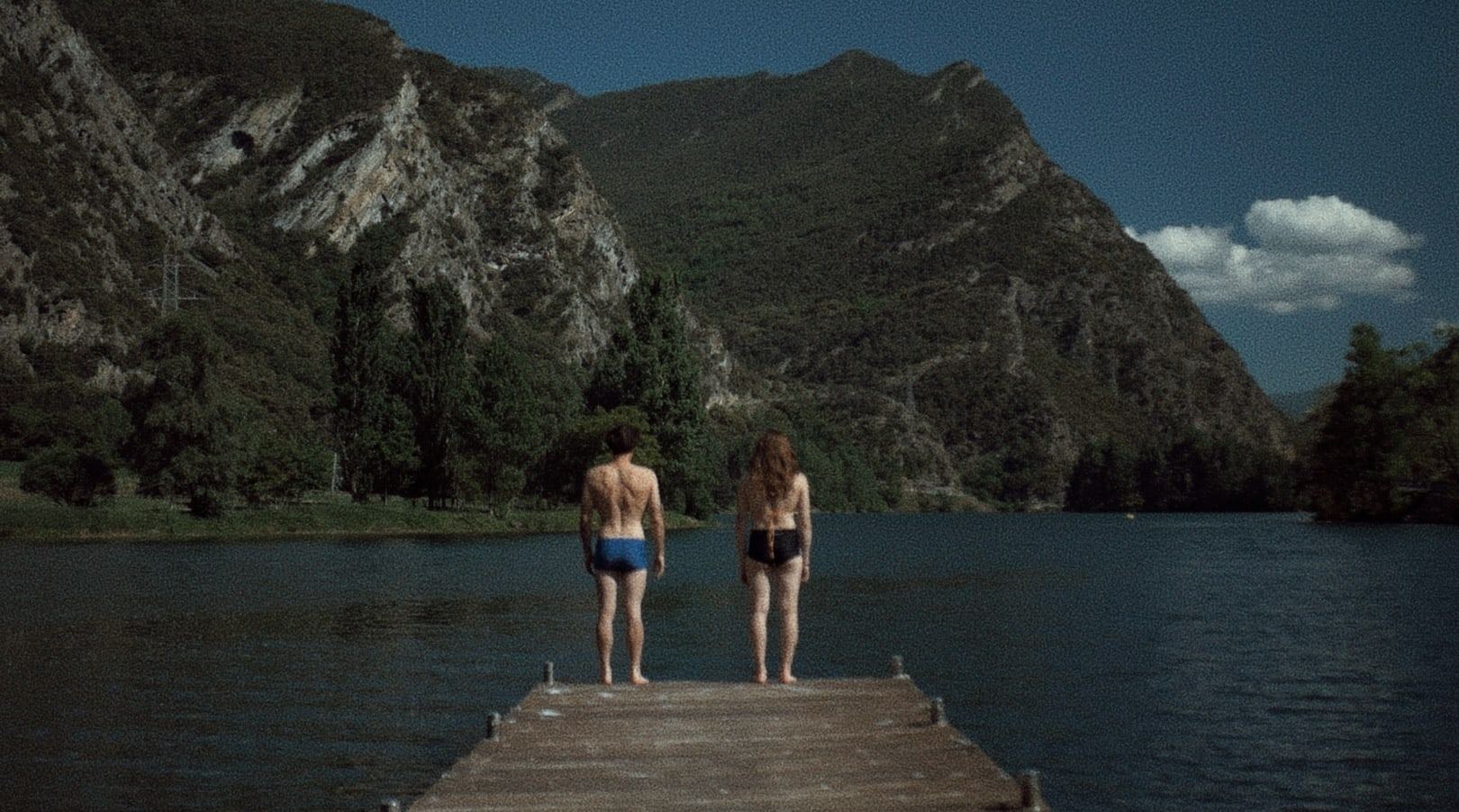 Two people standing on a jetty facing a lake and mountains