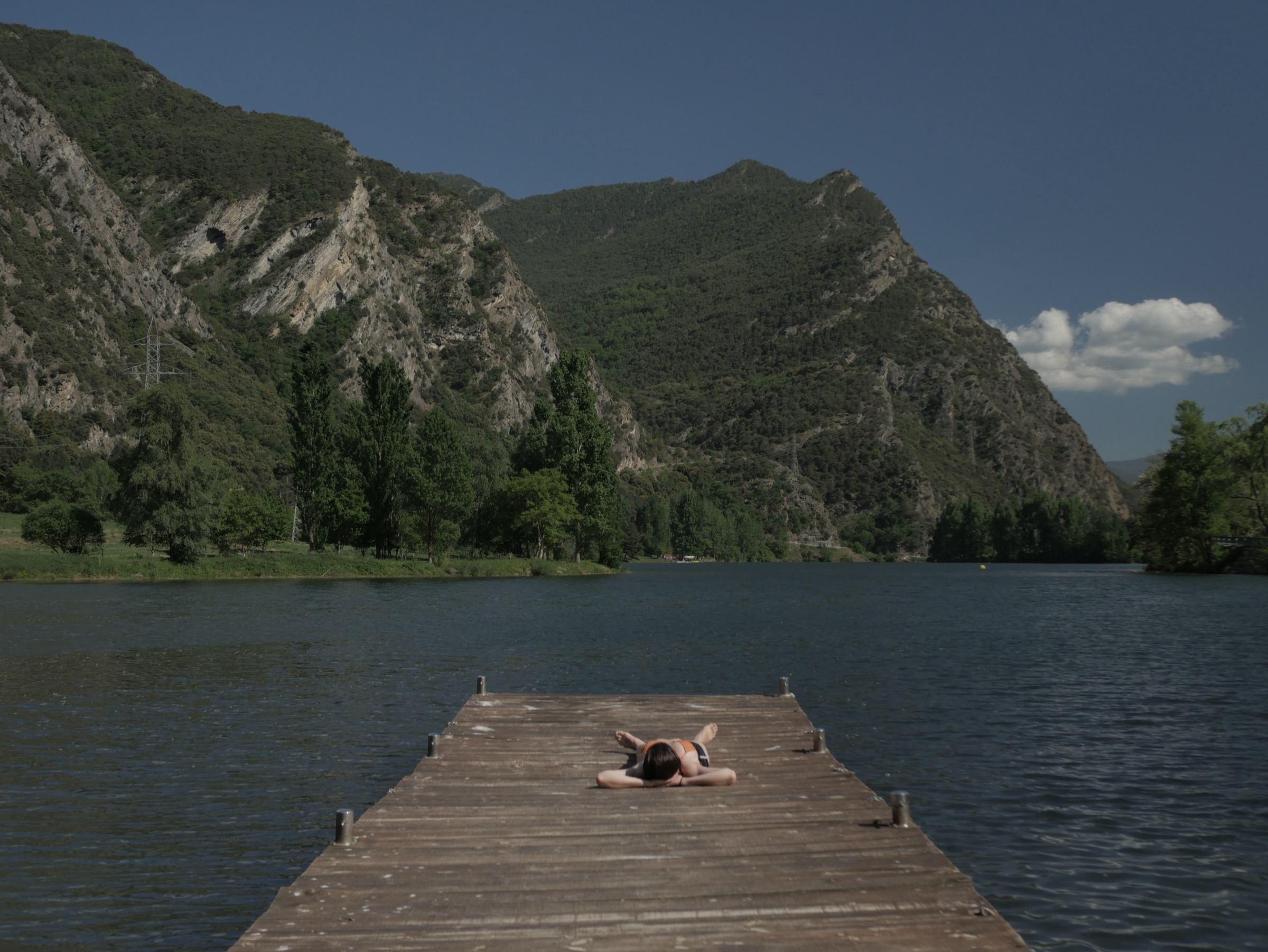 Person lying on a wooden jetty facing a lake and mountains