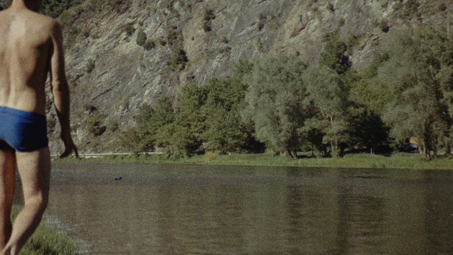 Man in blue swim trunks walking by a lake