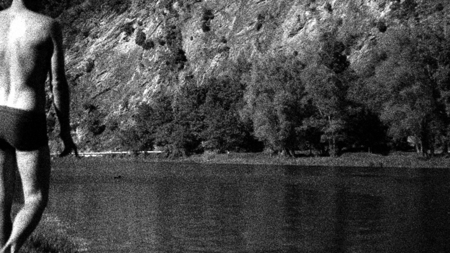 Hombre en traje de baño caminando junto a un lago, en blanco y negro